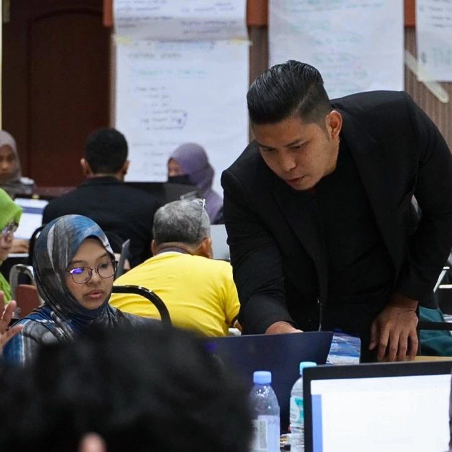 Suasana rapat semasa sesi amali dalam bengkel AI korporat di mana jurulatih membantu peserta menyelesaikan tugasan di komputer riba